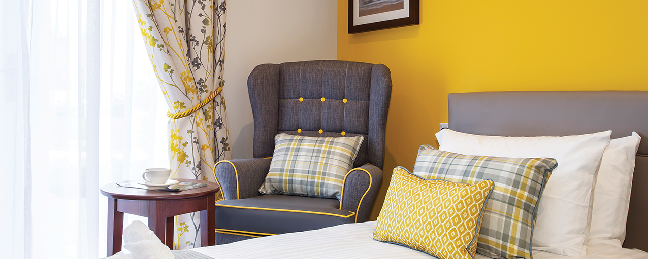 A close up of a bed and armchair in a yellow bedroom at a care home in Penrose Court Care Home